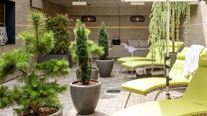 Een moderne, overdekte zithoek met potplanten, gele lounge fauteuils en een hangplant.