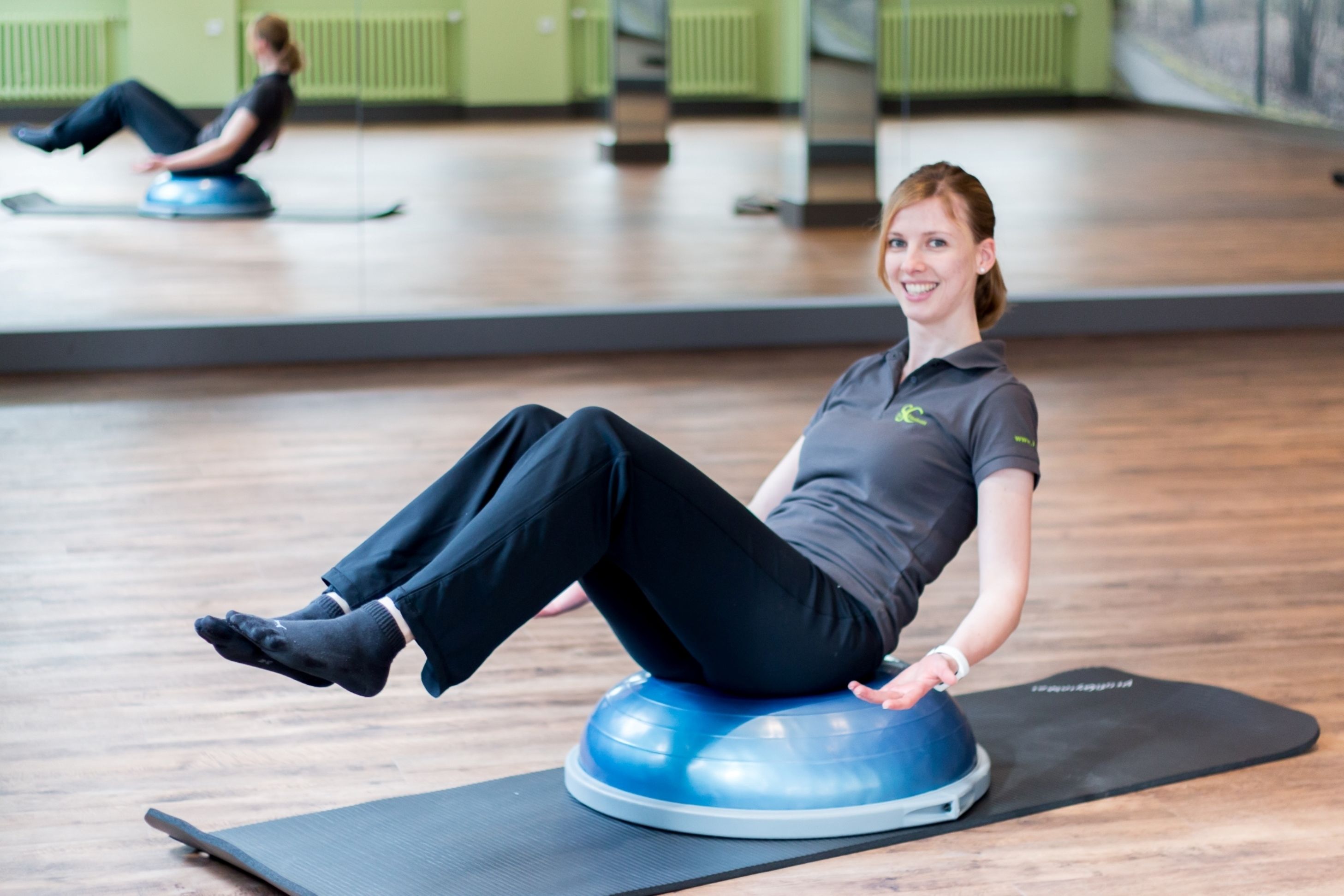 Eine Frau sitzt auf einem blauen Trainingsball in einem Fitnessstudio mit Holzboden und grünen Wänden.