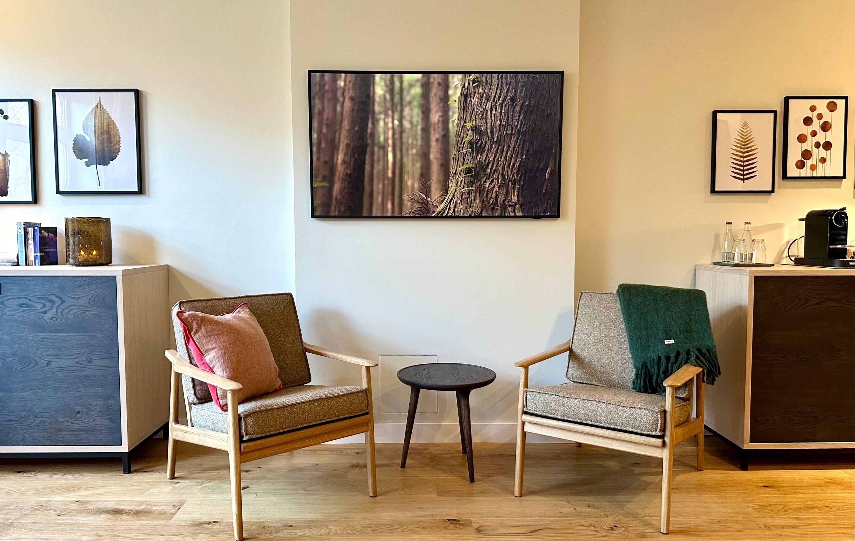 Ein modernes Wohnzimmer mit zwei Holzstühlen, einem kleinen runden Tisch und einem großen gerahmten Waldfoto an der Wand.