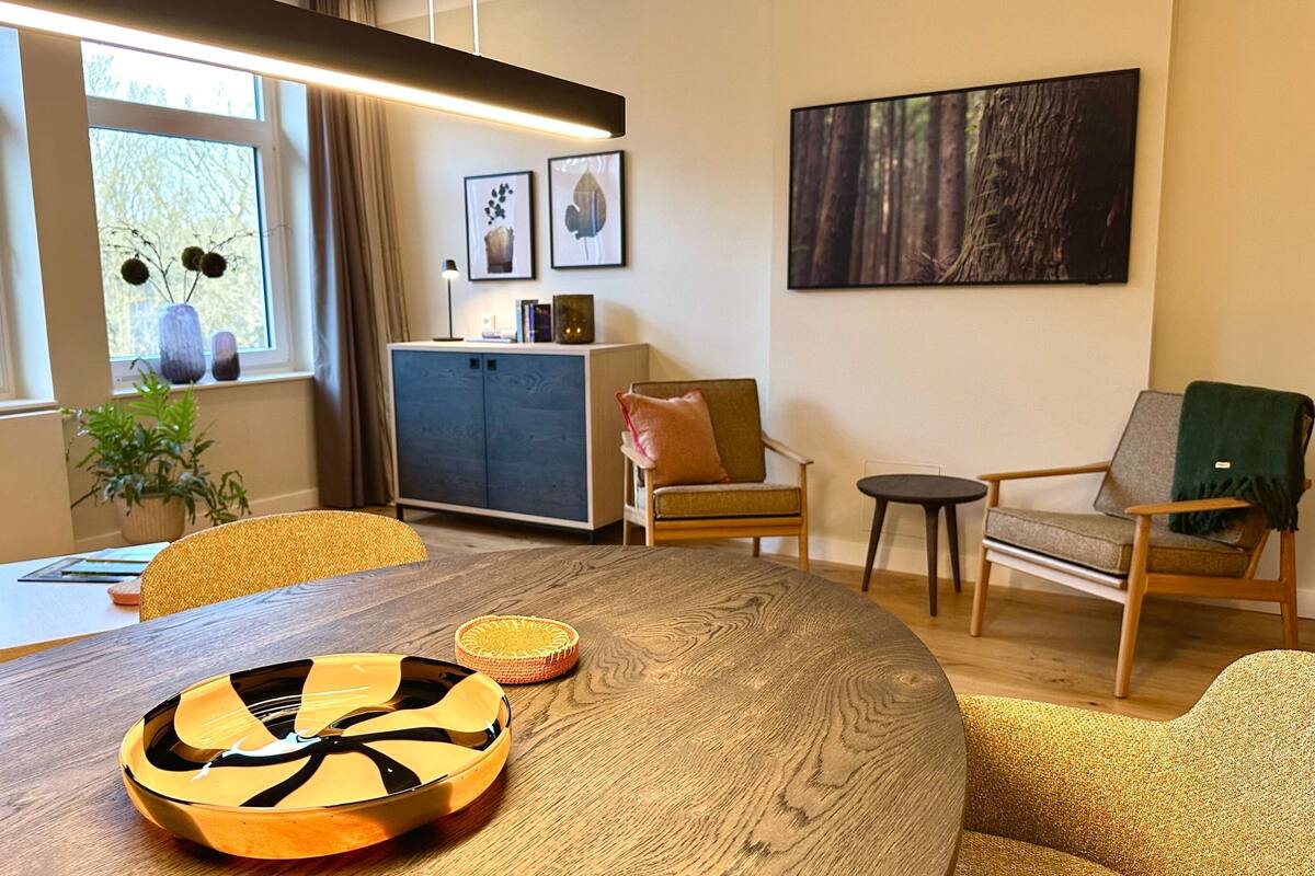 Een moderne woonkamer met een ronde houten tafel, een bank en twee stoelen. Er staat een kast met decoratieve objecten erop, een raam met uitzicht op bomen, en een schilderij aan de muur.