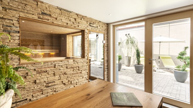 Een modern interieur met een stenen muur, een houten tafel en grote ramen met uitzicht op een buitenruimte met potplanten en een zithoek.