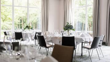A dining area with tables set for a meal, large windows with curtains, and a view of greenery outside.