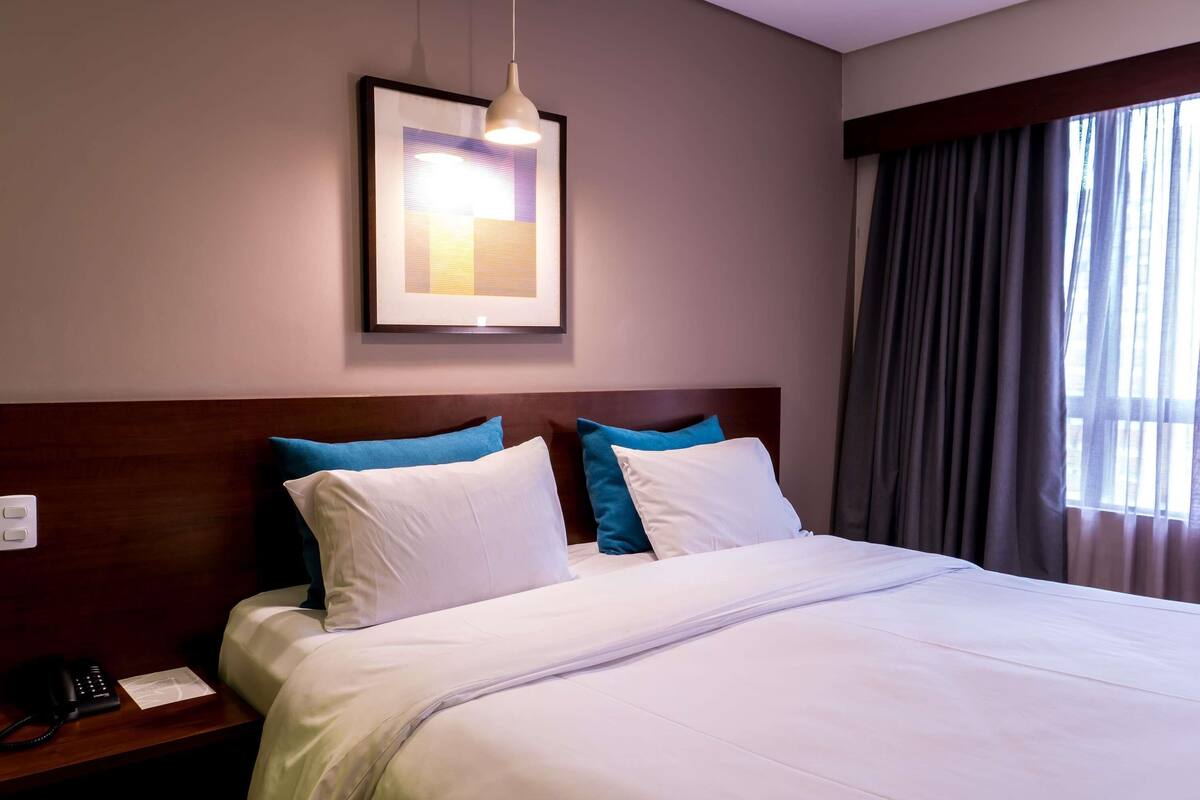 Una habitación de hotel con cama, mesita de noche, teléfono, un cuadro en la pared y ventana con cortinas.