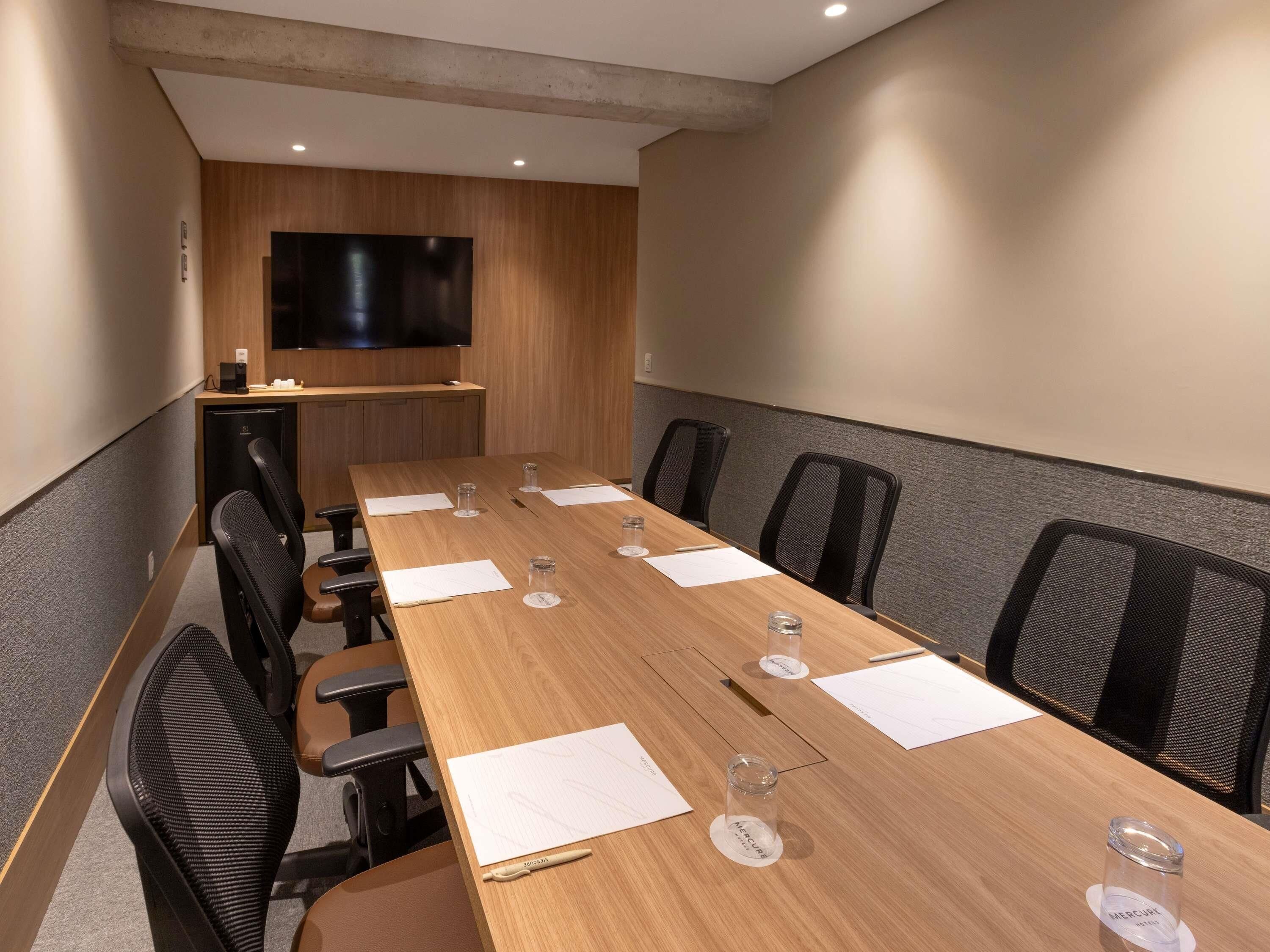 Un salón de conferencias con una larga mesa, sillas, un televisor de pantalla plana y una pequeña área de cocina.
