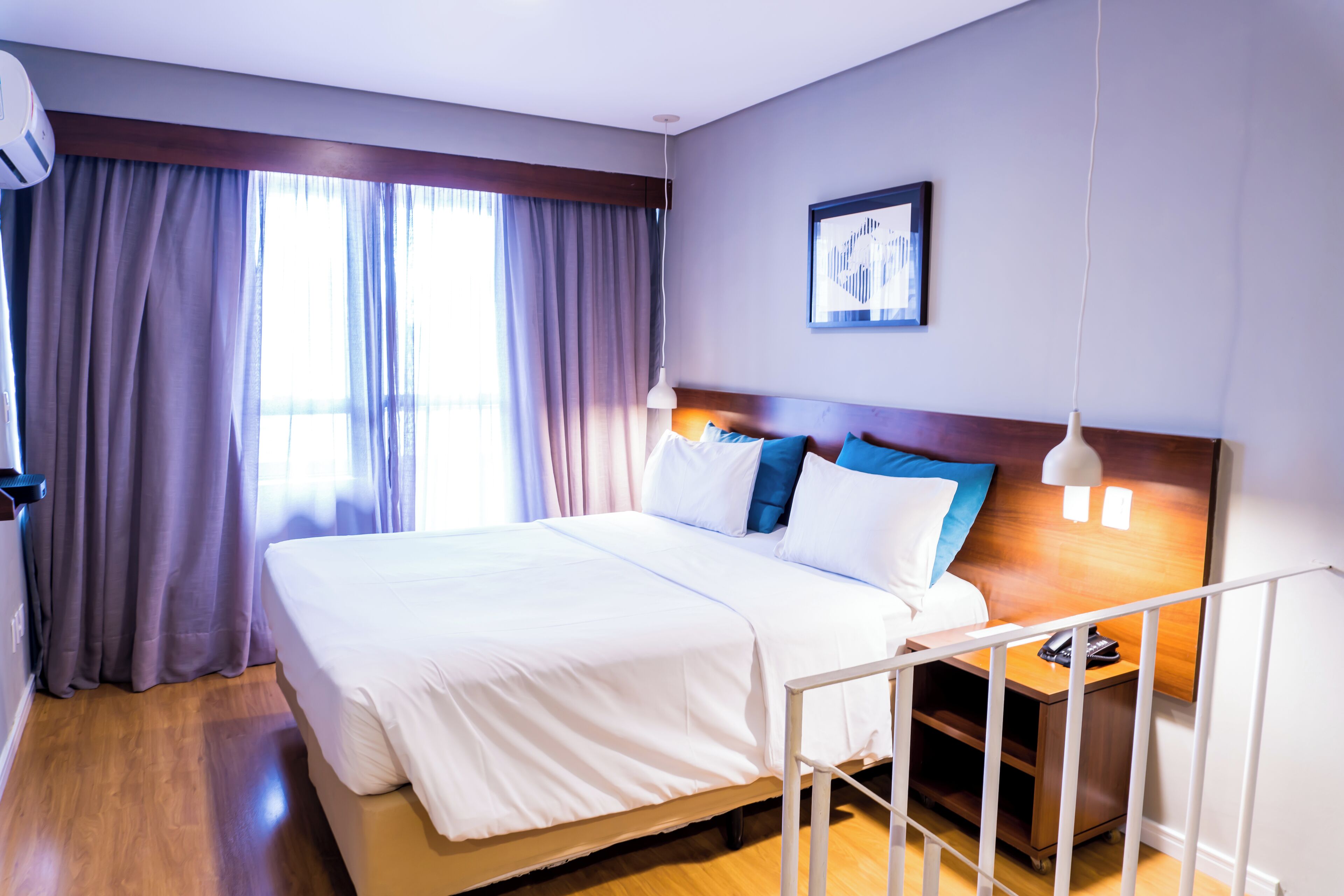 Una habitación de hotel con cama, mesita de noche, lámpara y ventana con cortinas.