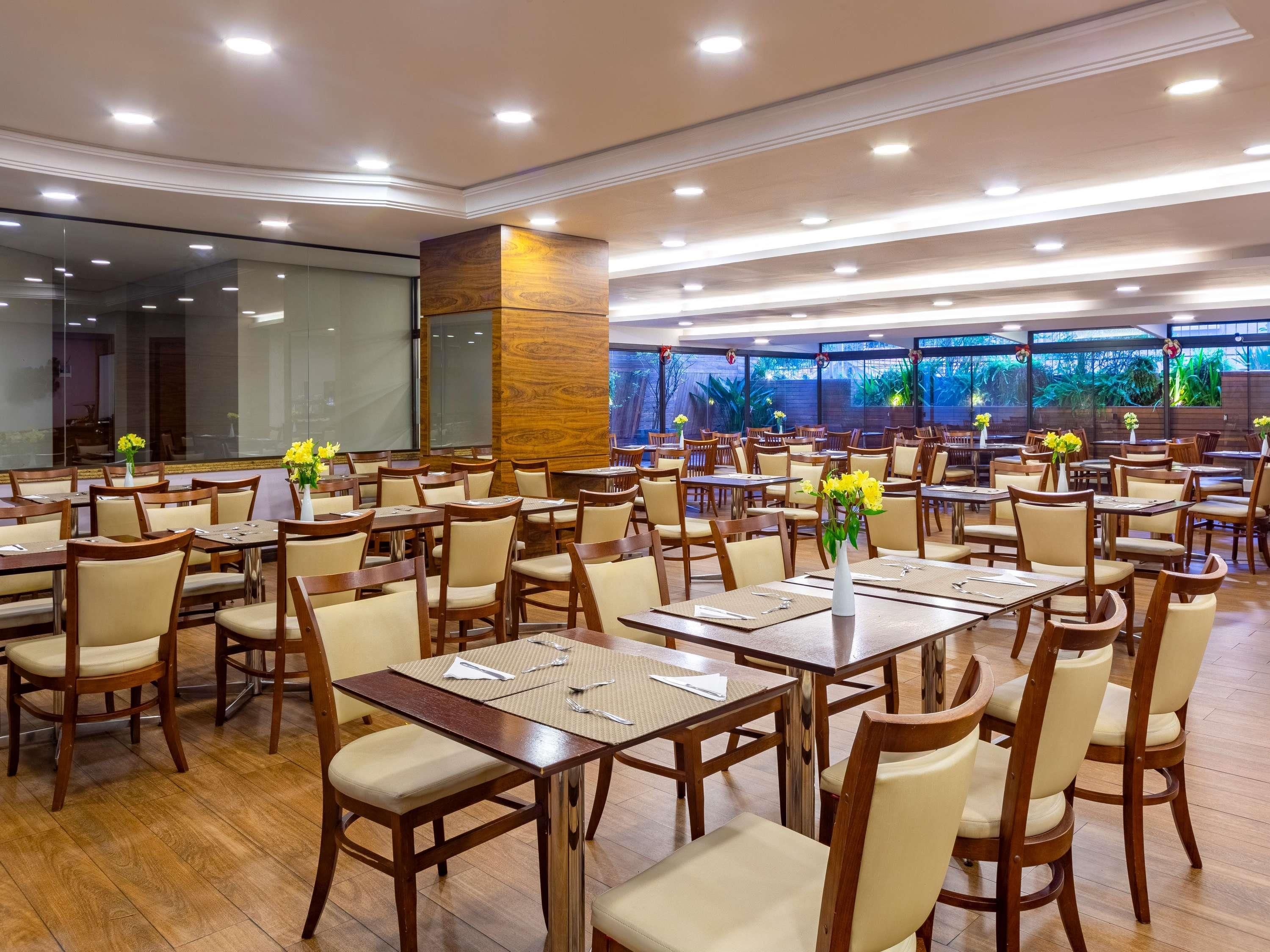 A spacious dining area with multiple tables and chairs, large windows with a view of greenery, and a modern ceiling design.