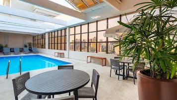 Una piscina interior con sillones de descanso y una mesa, rodeada de amplias ventanas y una planta en maceta.