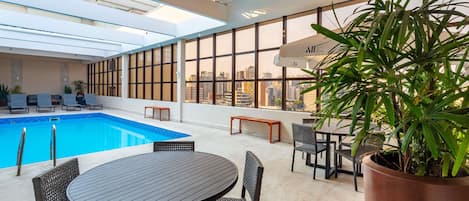 An indoor swimming pool with lounge chairs and a table, surrounded by large windows and a potted plant.