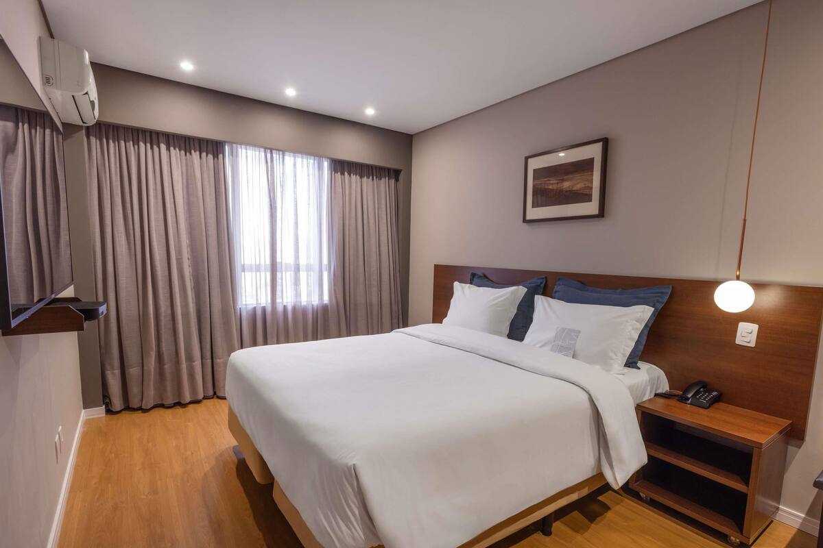 Una habitación de hotel con una cama grande, una mesita de noche con teléfono, aire acondicionado instalado en la pared y una ventana con cortinas.