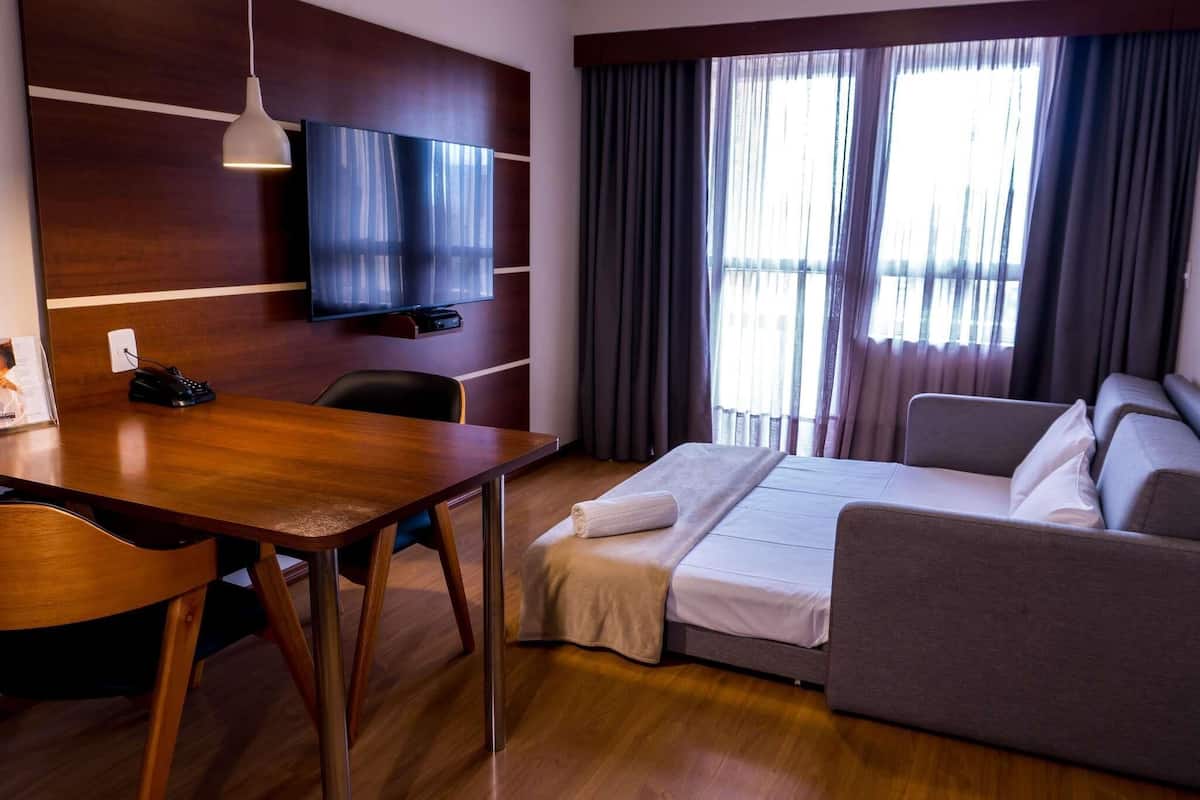 Et hotelværelse med et træskrivebord, et fjernsyn ophængt på væggen, en seng med hvid dyne, en grå sofa og et stort vindue med gardiner.