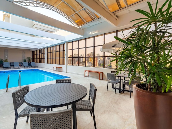 Une piscine intérieure dotée de chaises longues et d’une table, entourée de grandes fenêtres et agrémentée d’une plante en pot.