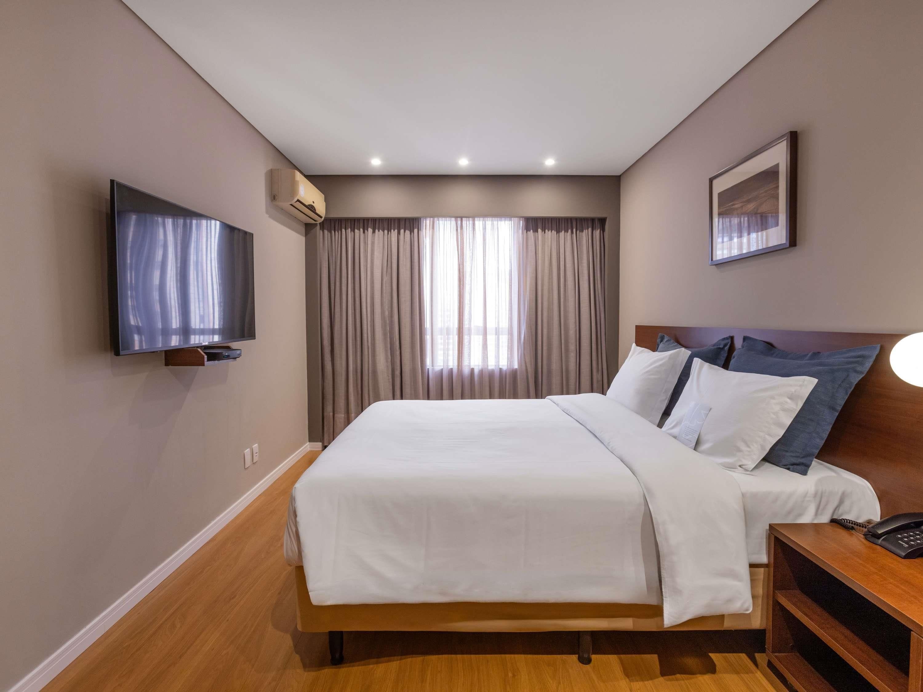 A hotel room with a bed, a TV, an air conditioner, a bedside table with a phone, and a painting on the wall.