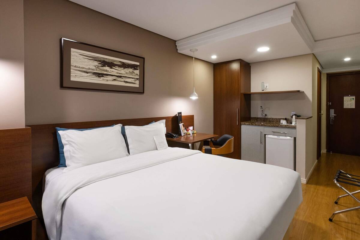 Una habitación de hotel con cama, mesita de noche, lámpara y una zona de kitchenette.