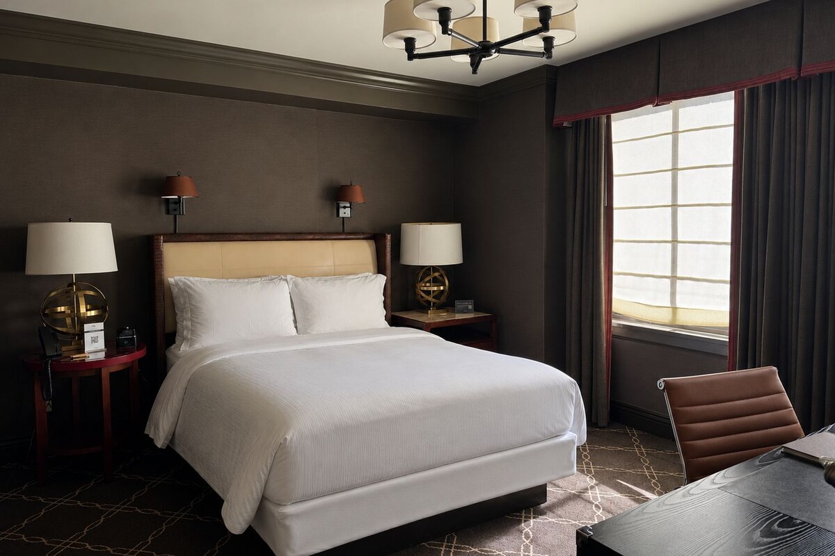 A hotel room with a large bed, two bedside tables with lamps, a chandelier, a window with curtains, and a table with a chair.