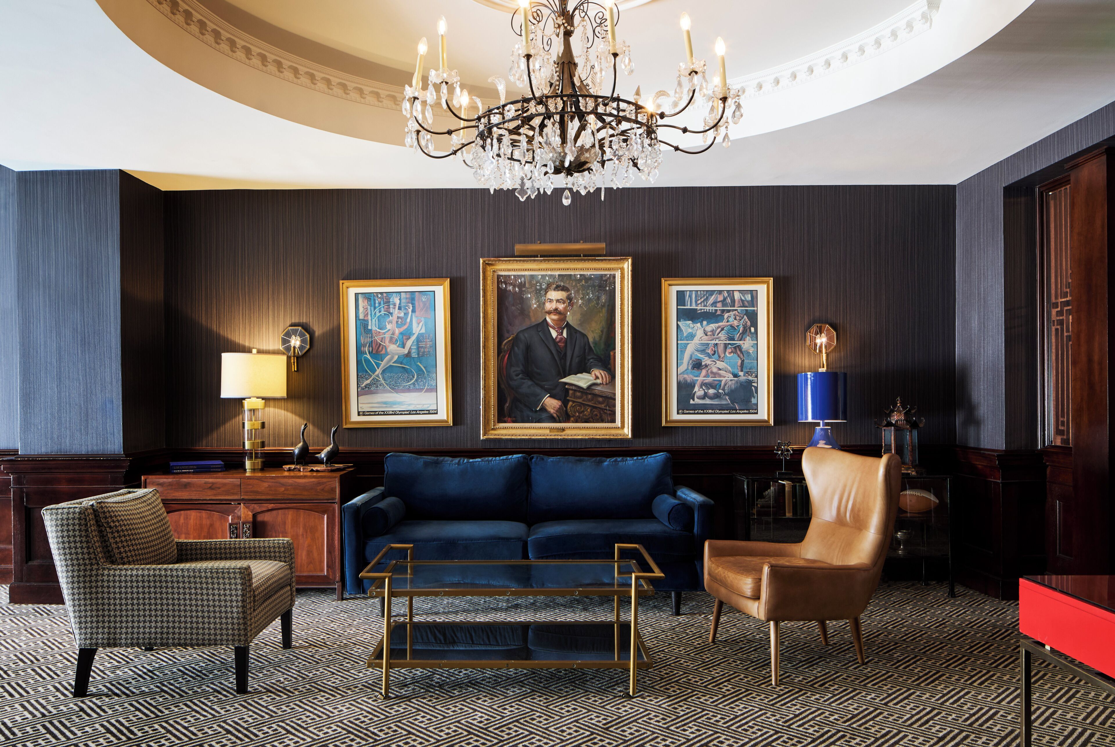 Een statige kamer met een kroonluchter, een blauwe bank, een bruine leren fauteuil en twee ingelijste kunstwerken aan de muur.