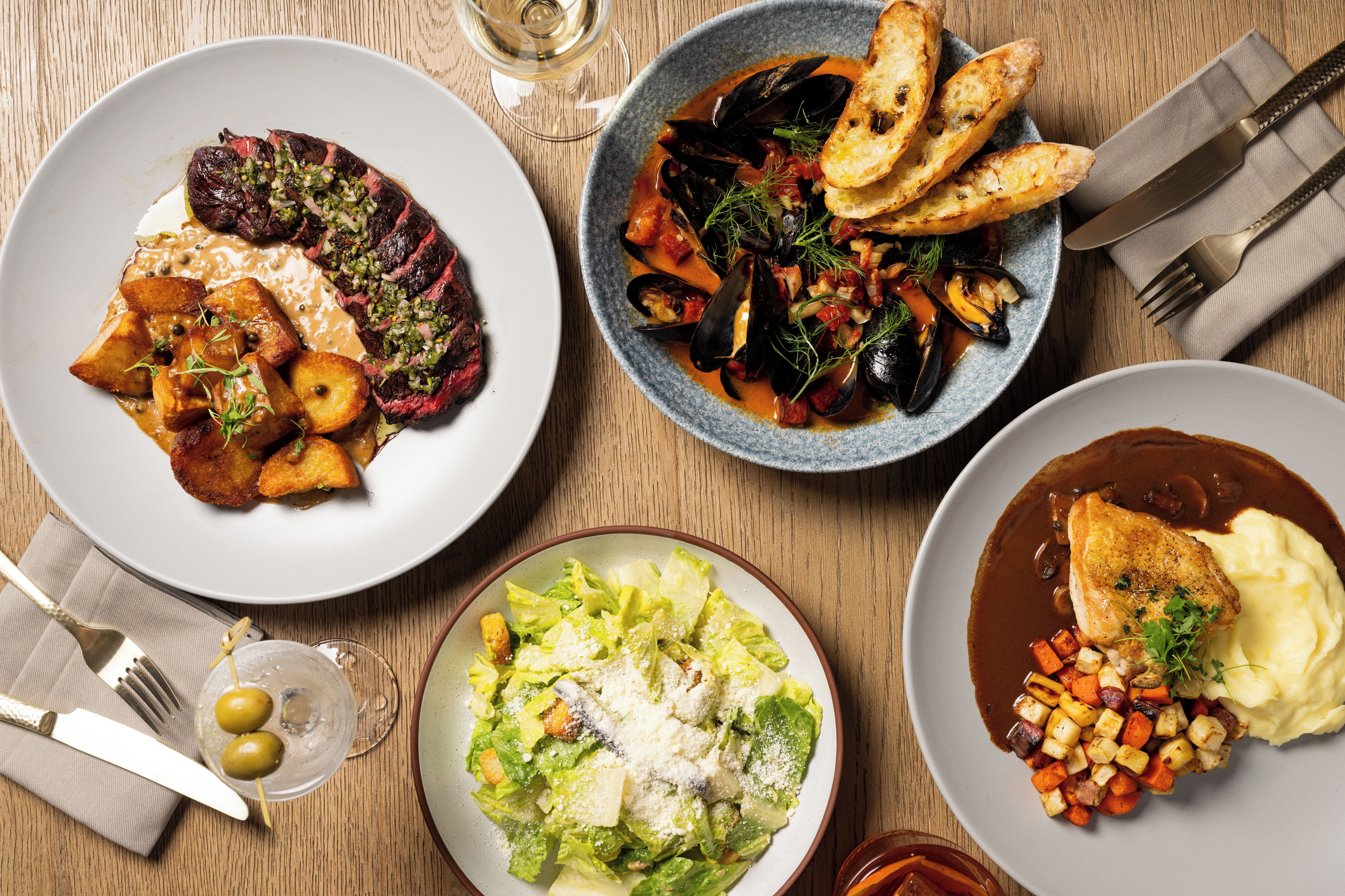 A table set with multiple dishes including steak, mussels, and a salad, with cutlery and a glass of wine.