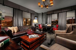 A hotel room with a leather sofa, a wooden coffee table, a red lamp, and a view of the cityscape through large windows.