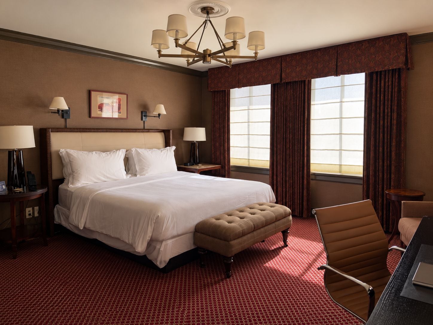 Habitación de hotel con una cama grande, un escritorio con silla, una araña y un ventanal con cortinas.