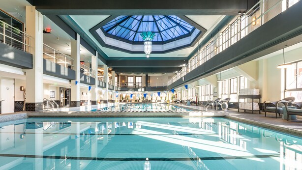 Una amplia piscina interior con techo de tragaluces, varios niveles con balcones y una zona de estar con sillas y mesas.