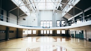 Una amplia cancha de baloncesto cubierta con techo alto y tragaluces, rodeada de paredes con ventanas y puertas.
