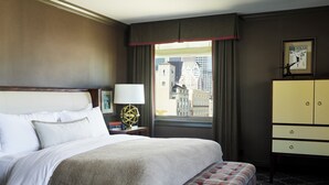 Habitación de hotel con una cama grande, una mesita de noche con lámpara, vista de la ciudad a través de una ventana y un cuadro en la pared.