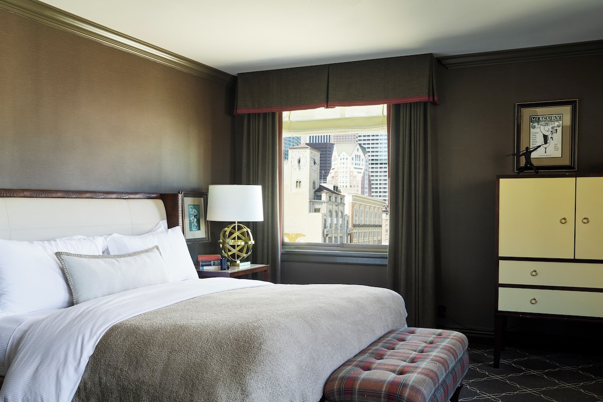 Habitación de hotel con una cama grande, una mesita de noche con lámpara, vista de la ciudad a través de una ventana y un cuadro en la pared.