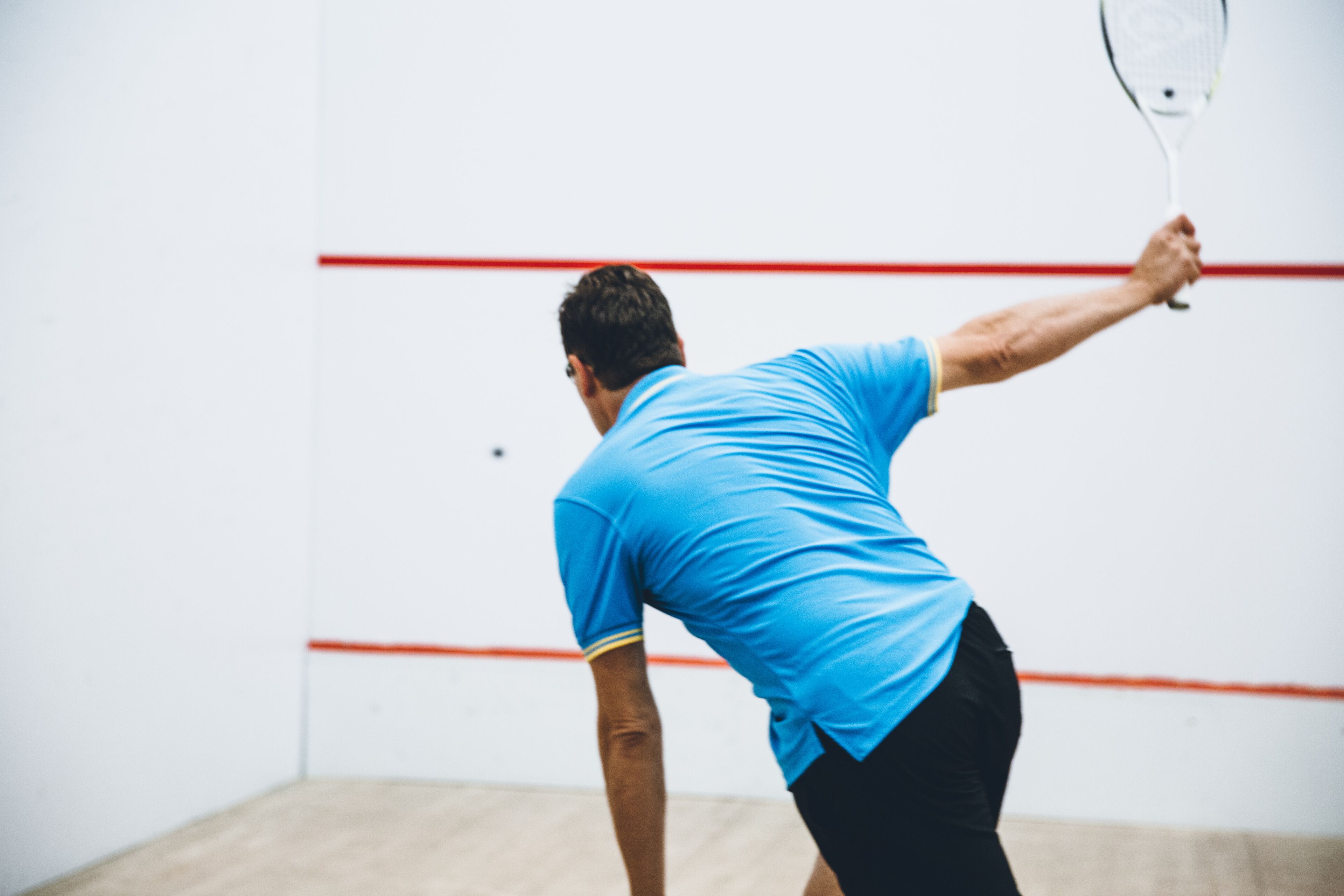 A squash player in a blue shirt and black shorts, holding a racket, in a room with a wooden floor and red lines on the wall.