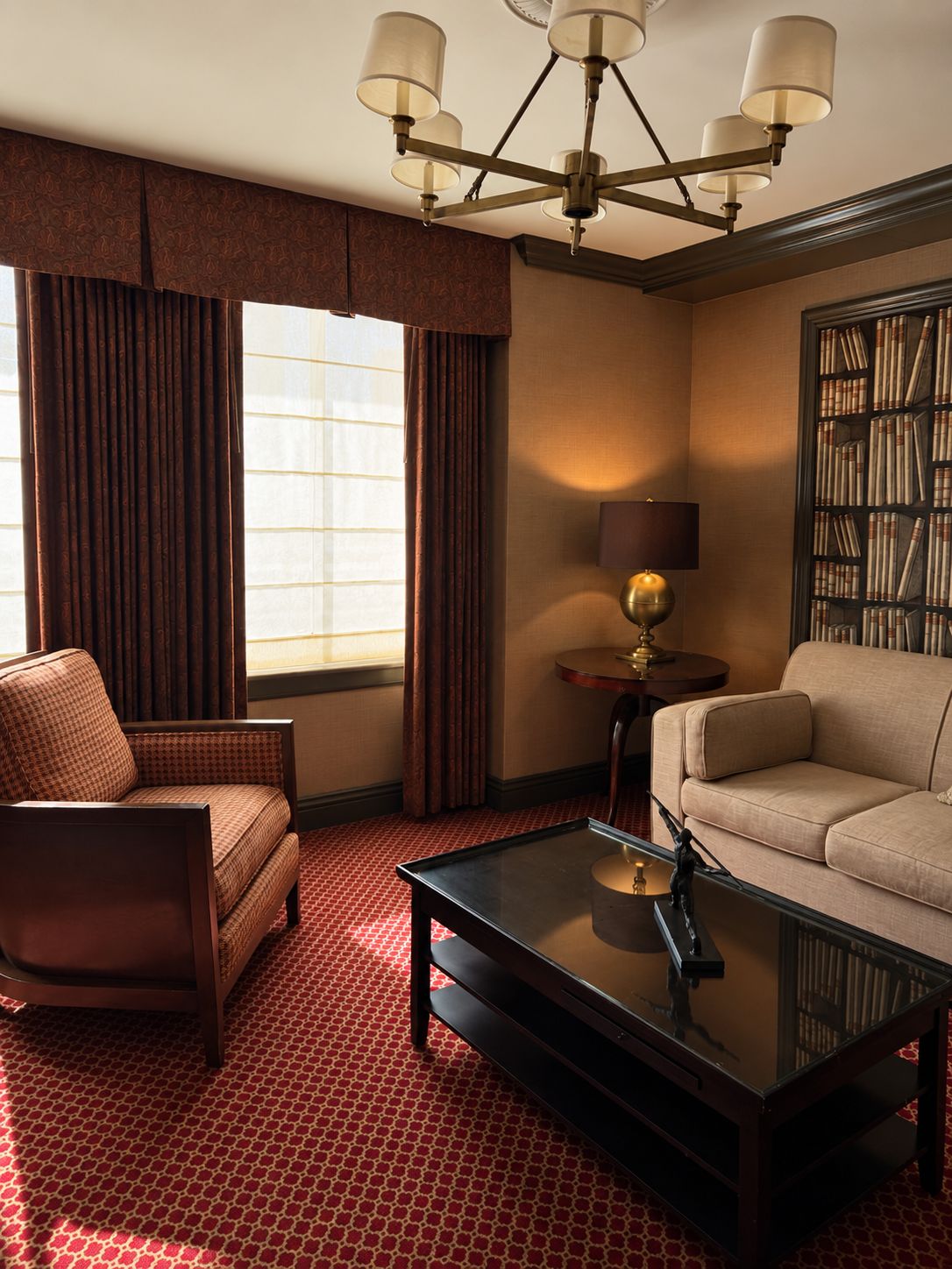 A hotel room with a sofa, armchair, and a coffee table. There is a bookshelf with books and a lamp on a side table.