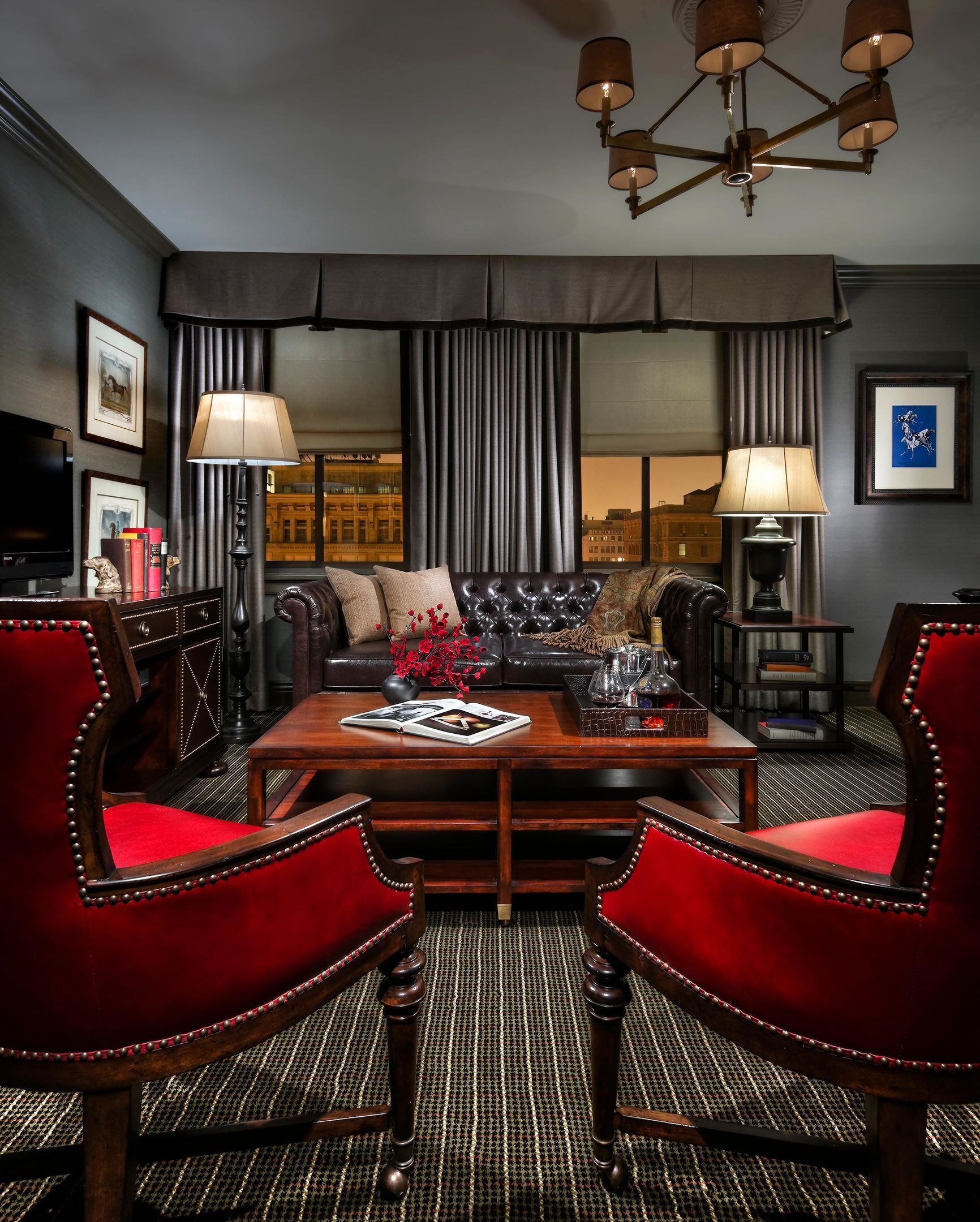 A room with a dark-colored sofa, a wooden coffee table, red upholstered chairs, a lamp, and a view of buildings through large windows.