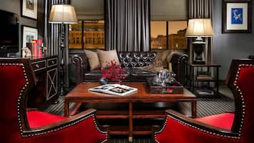 A room with a dark-colored sofa, a wooden coffee table, red upholstered chairs, a lamp, and a view of buildings through large windows.