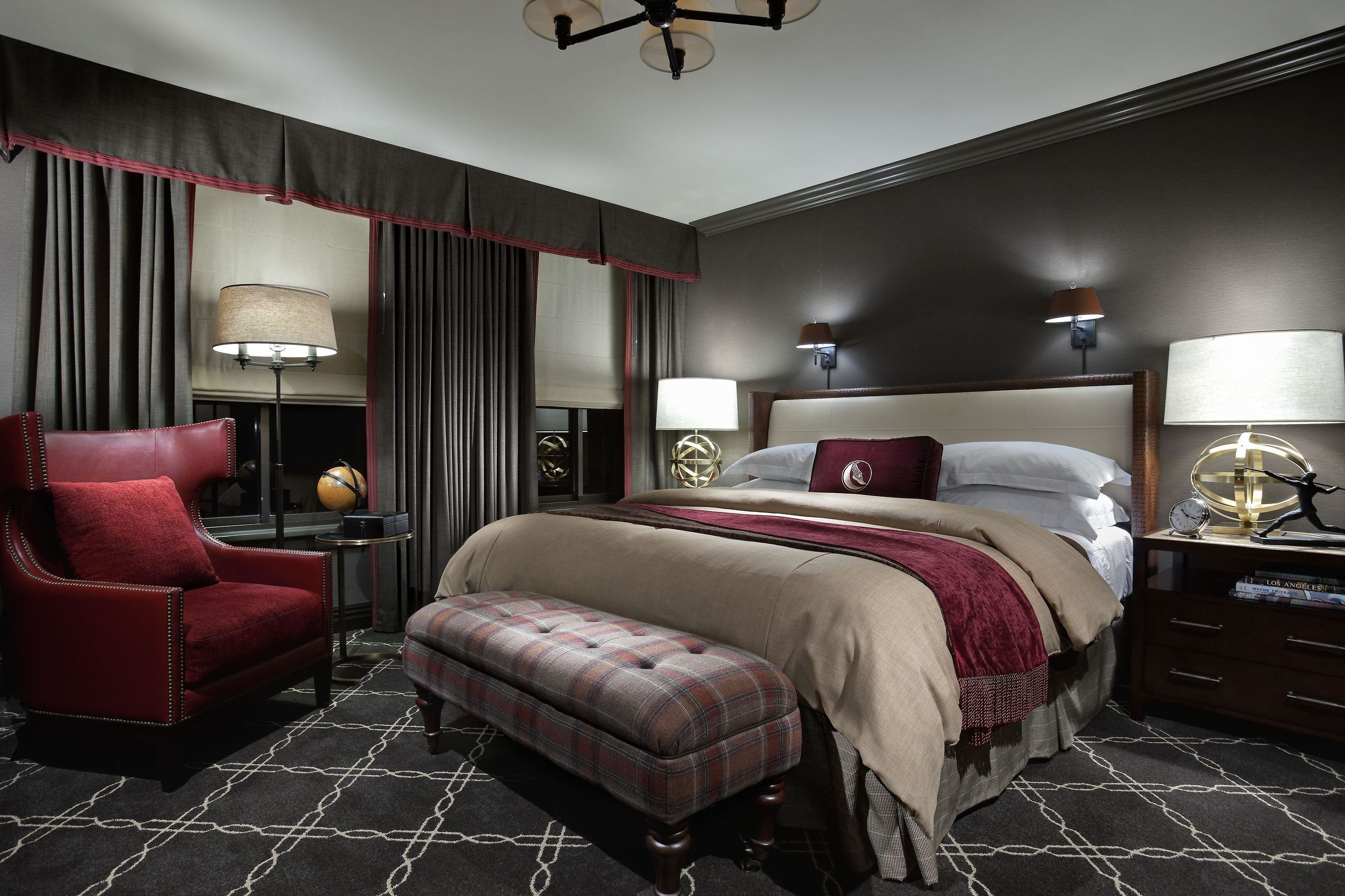 A hotel room with a large bed, a red armchair, a bench, a nightstand with a lamp, and a wall-mounted clock.