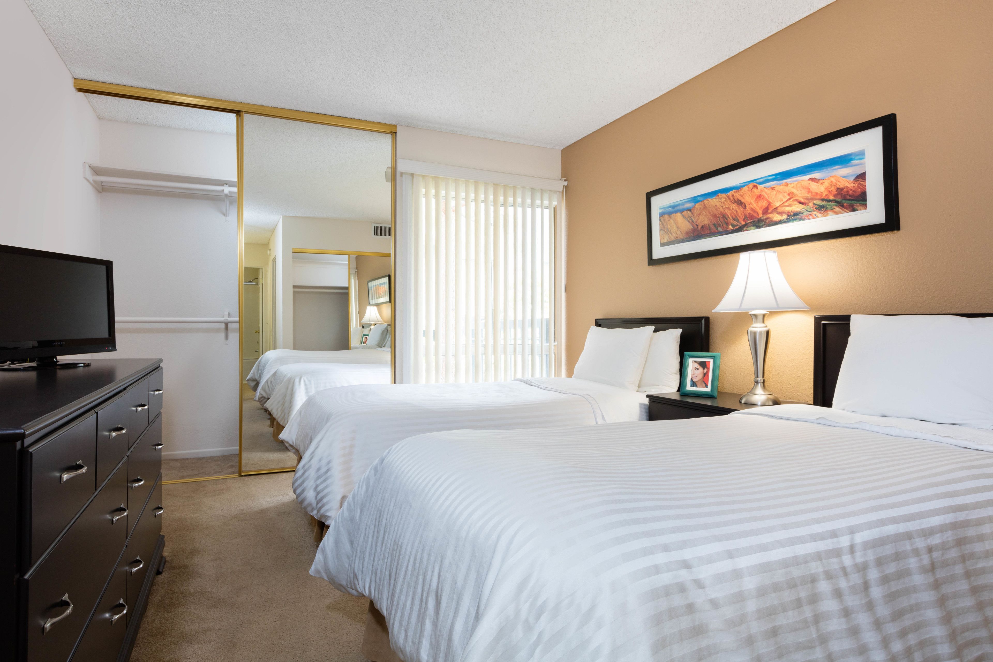 A hotel room with two beds, a TV, a dresser, a mirror, a painting, and a lamp.