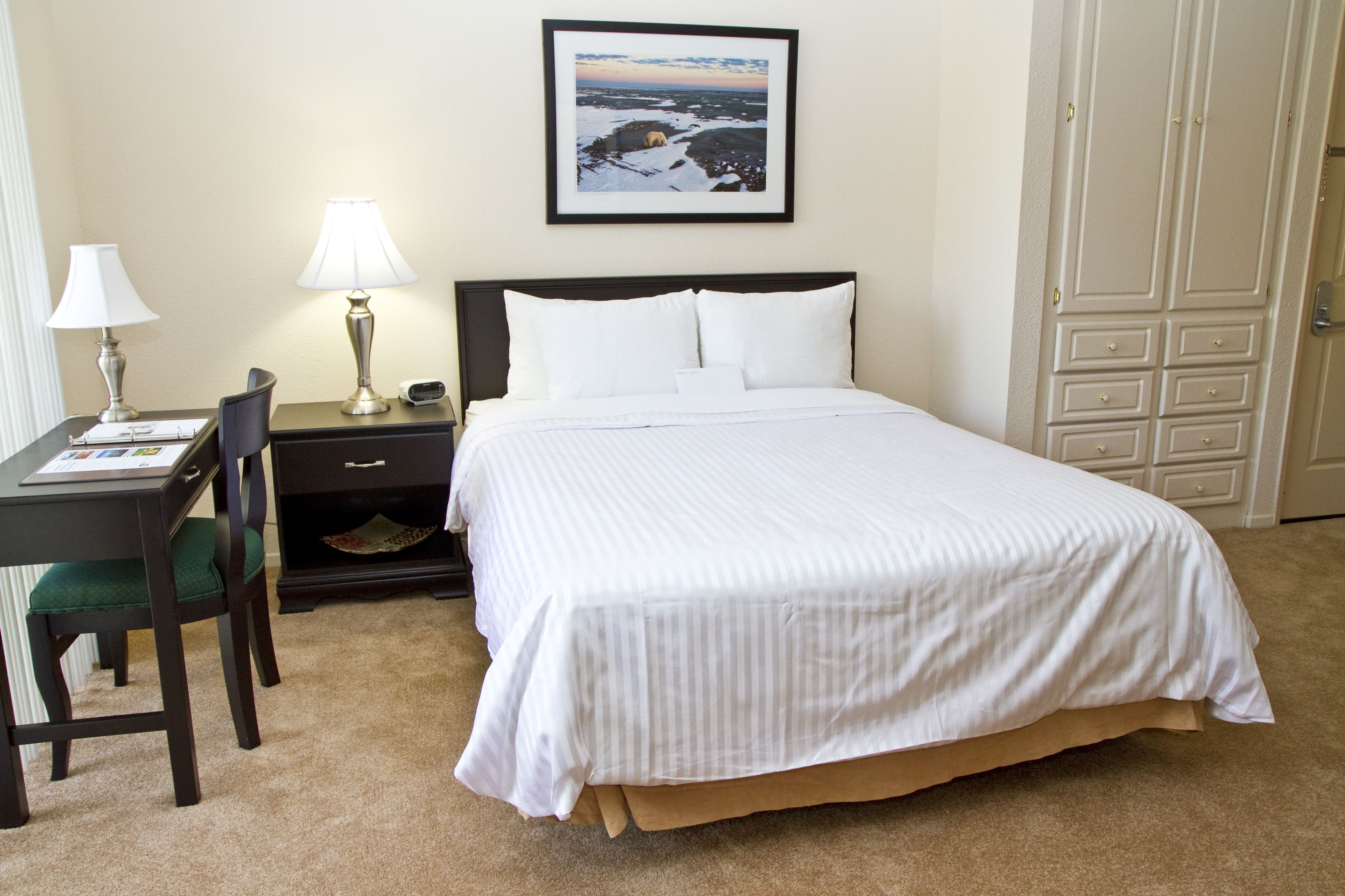 A neatly arranged hotel room with a bed, a desk with a lamp, a chair, and a framed picture on the wall.