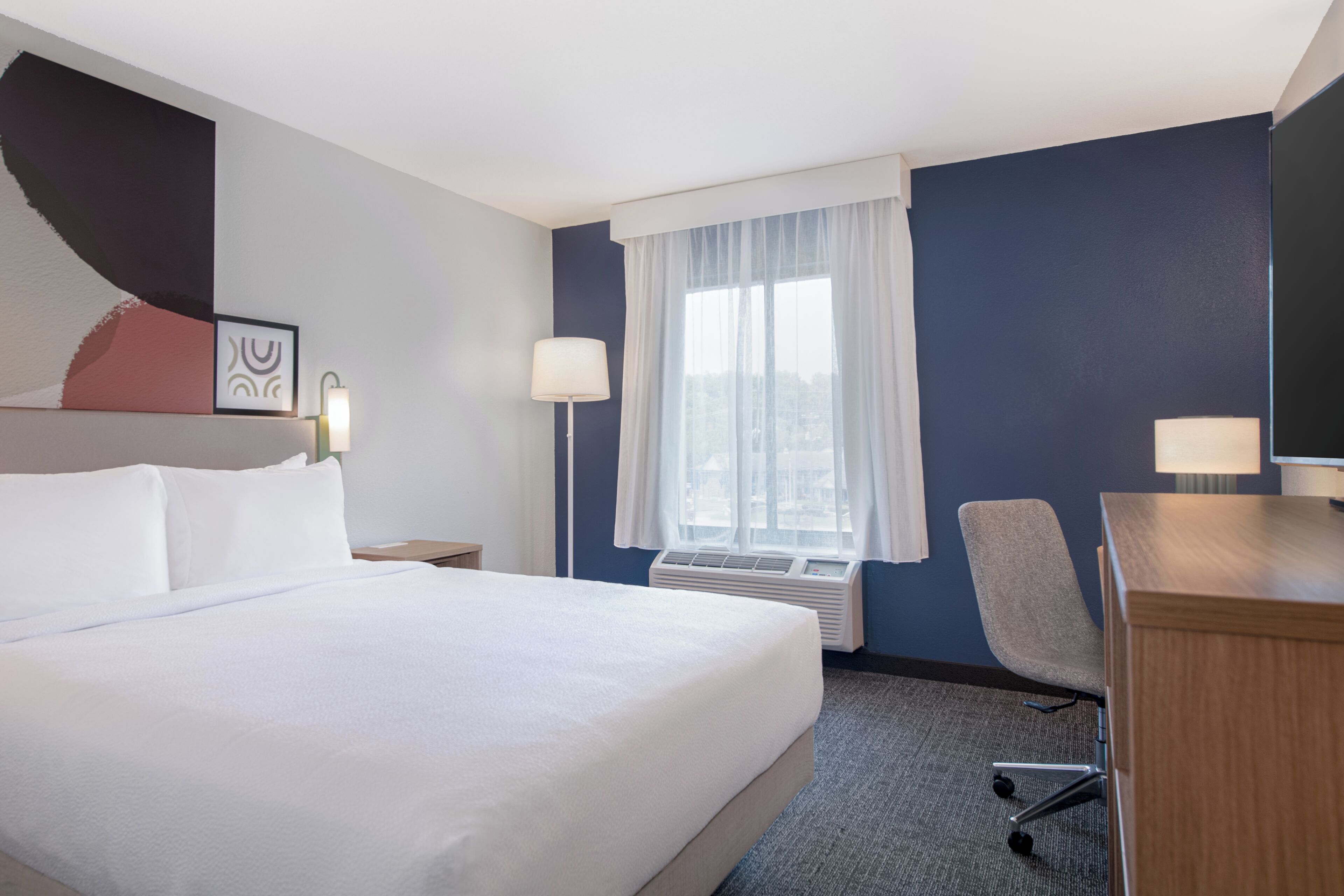 Habitación de hotel con una cama grande, un escritorio, una silla, un ventanal con cortinas y un aire acondicionado instalado en la pared.