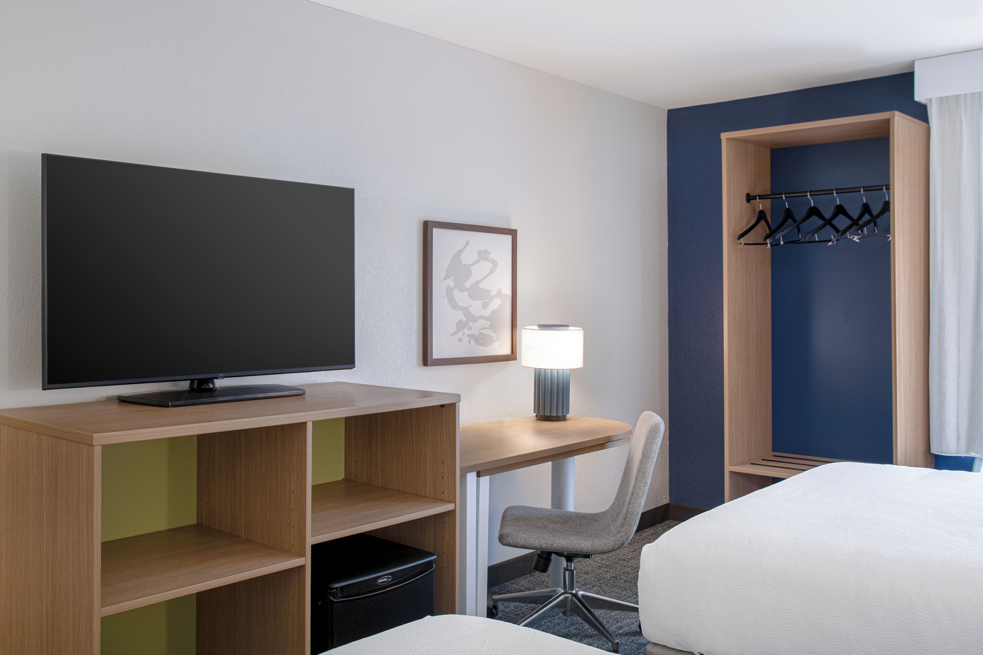 A hotel room with a flat-screen TV, a desk with a lamp, a chair, a bed, a coat rack, and a framed picture on the wall.