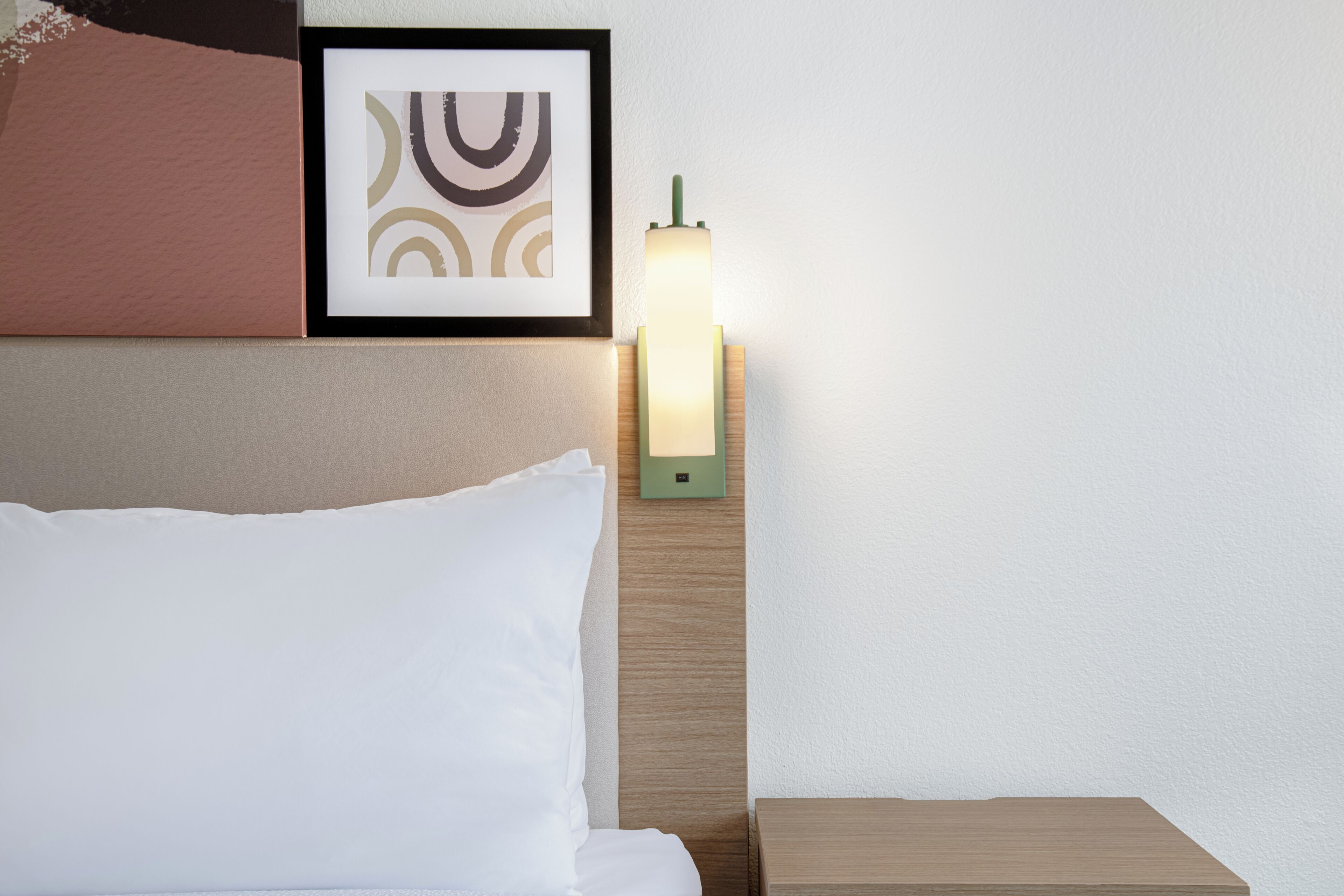 Un lit bien fait, avec des draps blancs, une œuvre d’art abstraite encadrée au-dessus, et une table de chevet sur laquelle se trouve une lampe verte.
