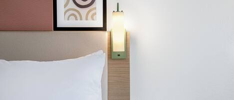 A neatly made bed with white sheets, a framed abstract artwork above it, and a bedside table with a green lamp.
