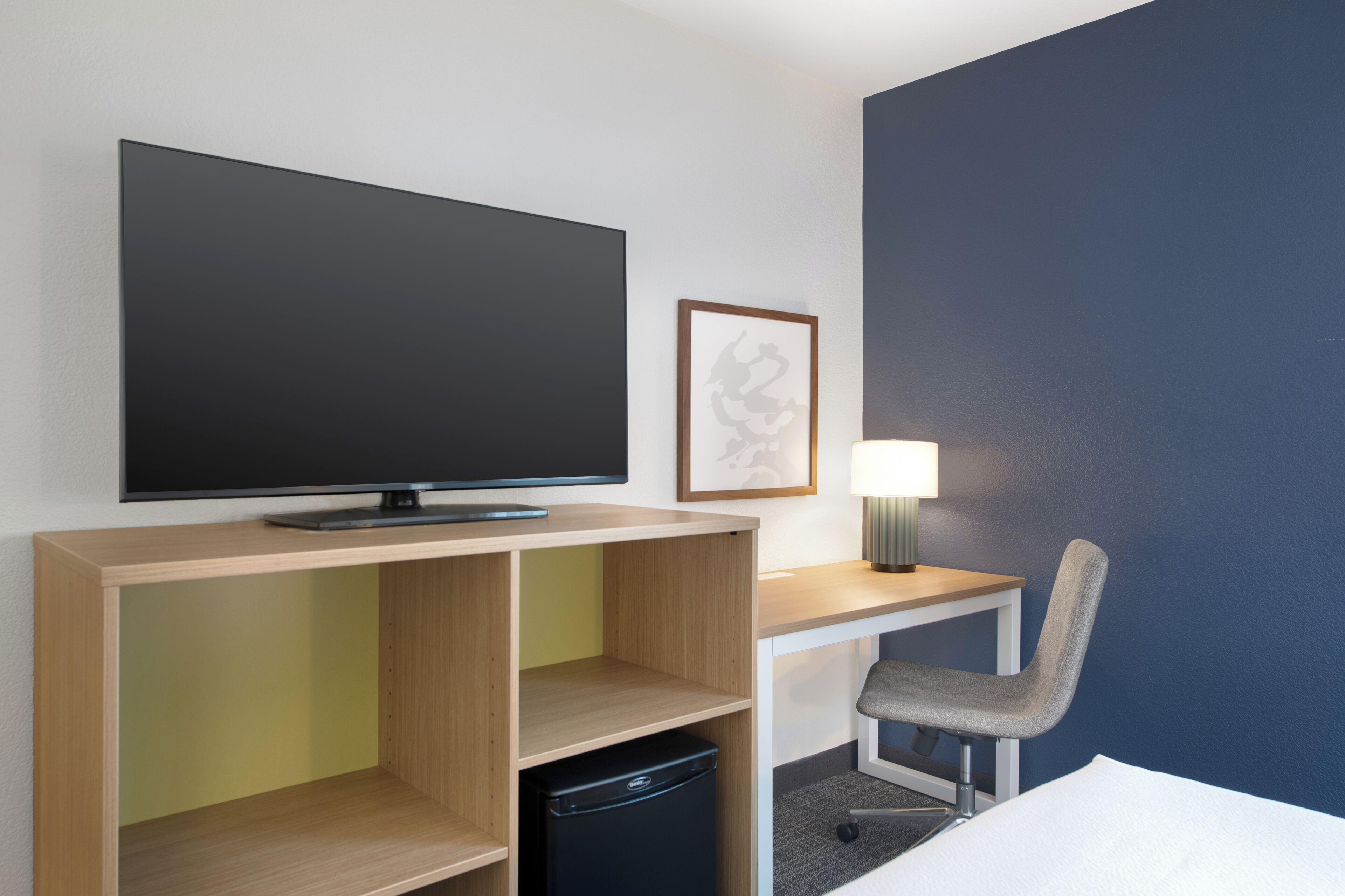 A hotel room with a flat-screen TV, a desk with a chair, a lamp, and a bed.