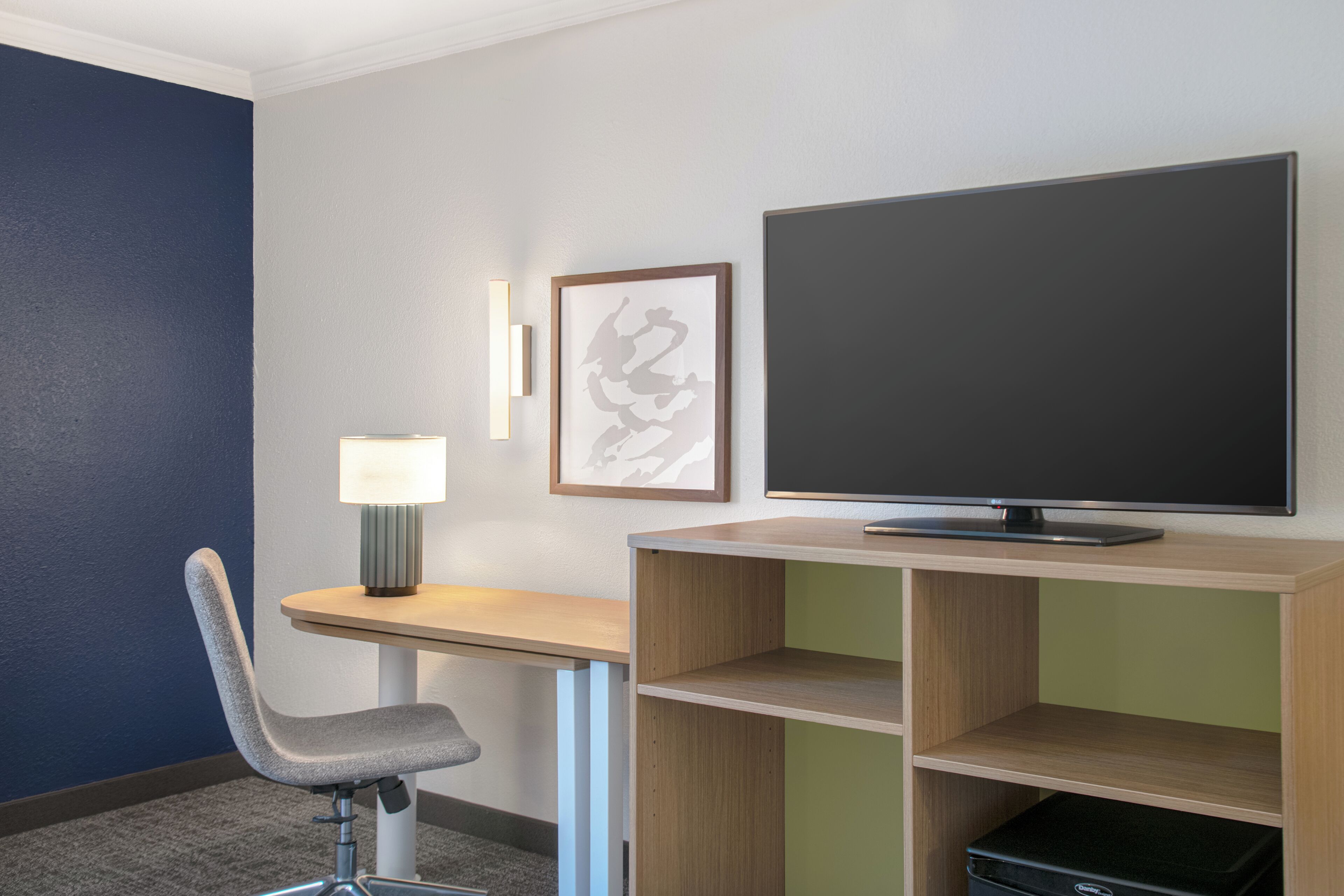 A hotel room with a desk, chair, TV, lamp, and artwork on the wall.
