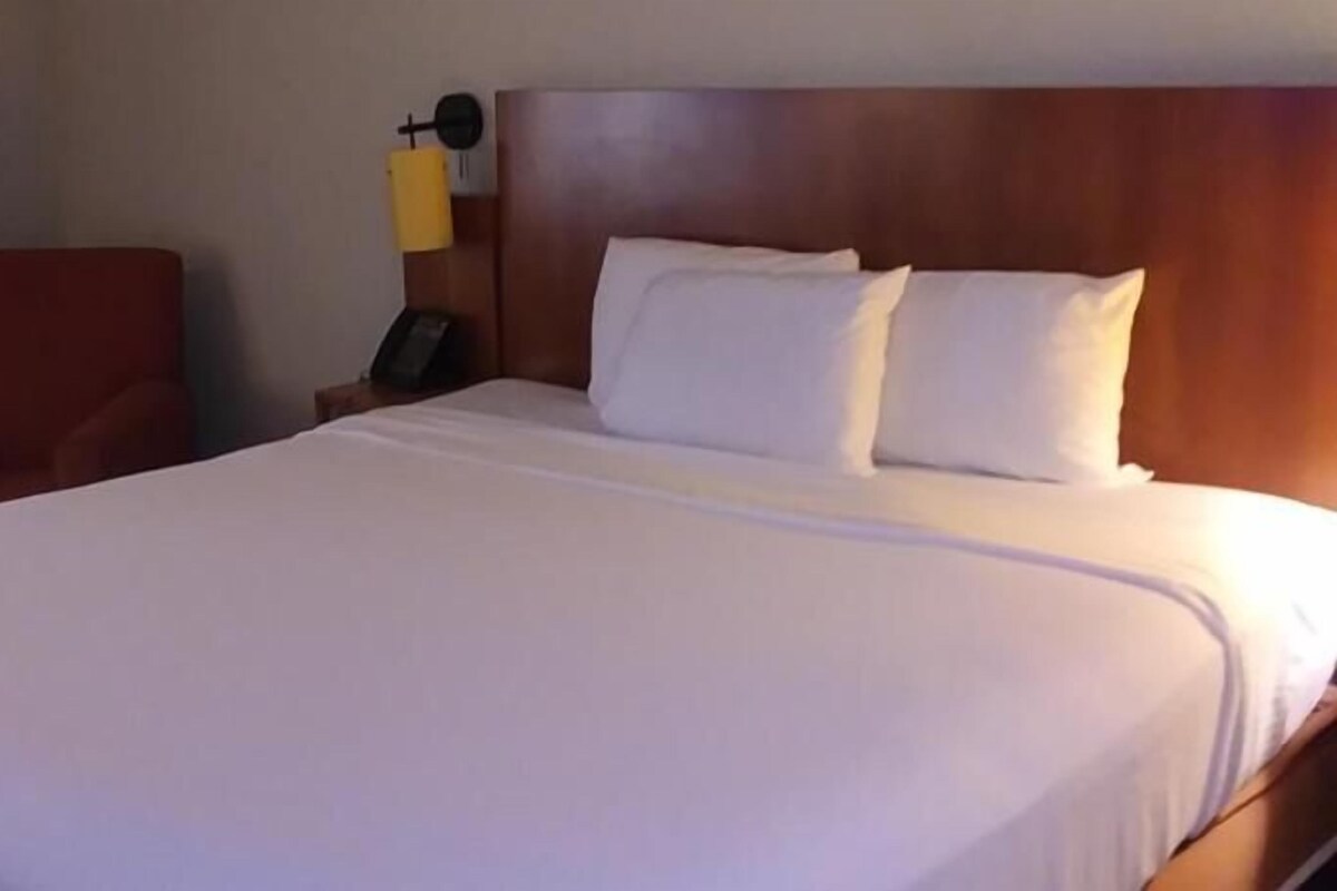 A hotel room with a large bed, a wooden headboard, a nightstand, a chair, and a wall-mounted lamp.