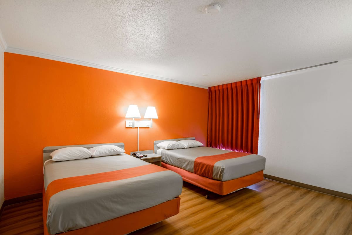 A hotel room with two beds, a nightstand, a lamp, and a window with curtains.