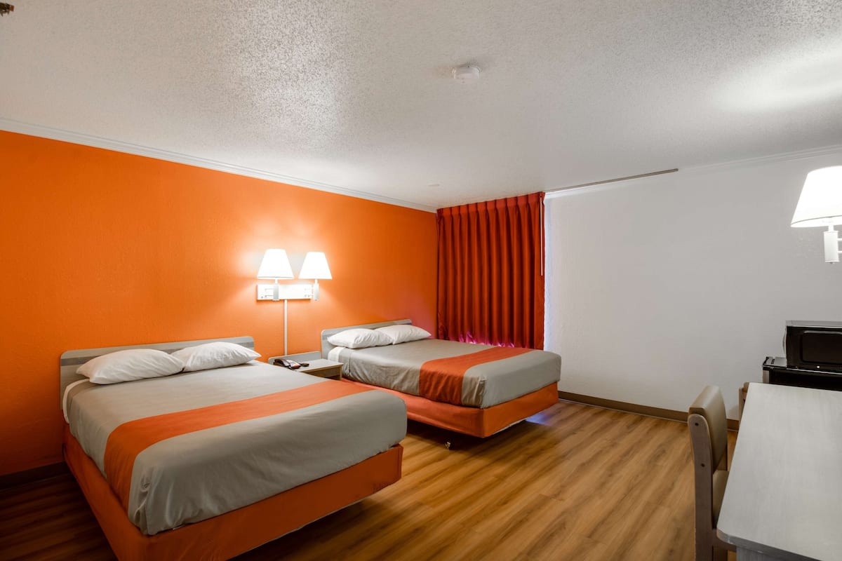 A hotel room with two beds, a desk, a chair, and a lamp.