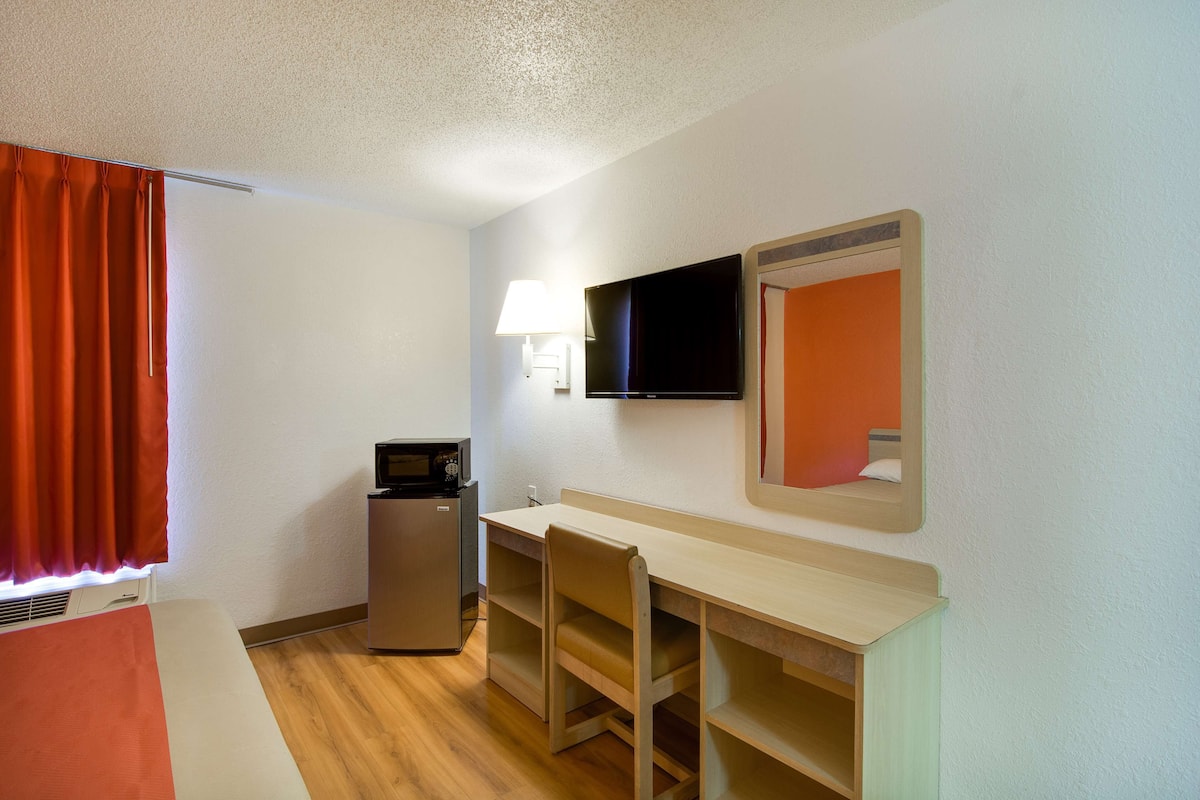 A hotel room with a bed, a desk, a chair, a television, a mirror, a lamp, and a small refrigerator.