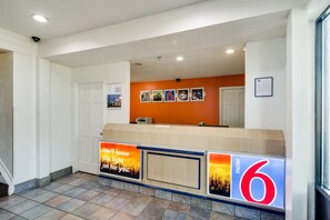 Un espace d’accueil d’hôtel avec un comptoir, une enseigne indiquant « Motel 6 », et un mur orné de tableaux.