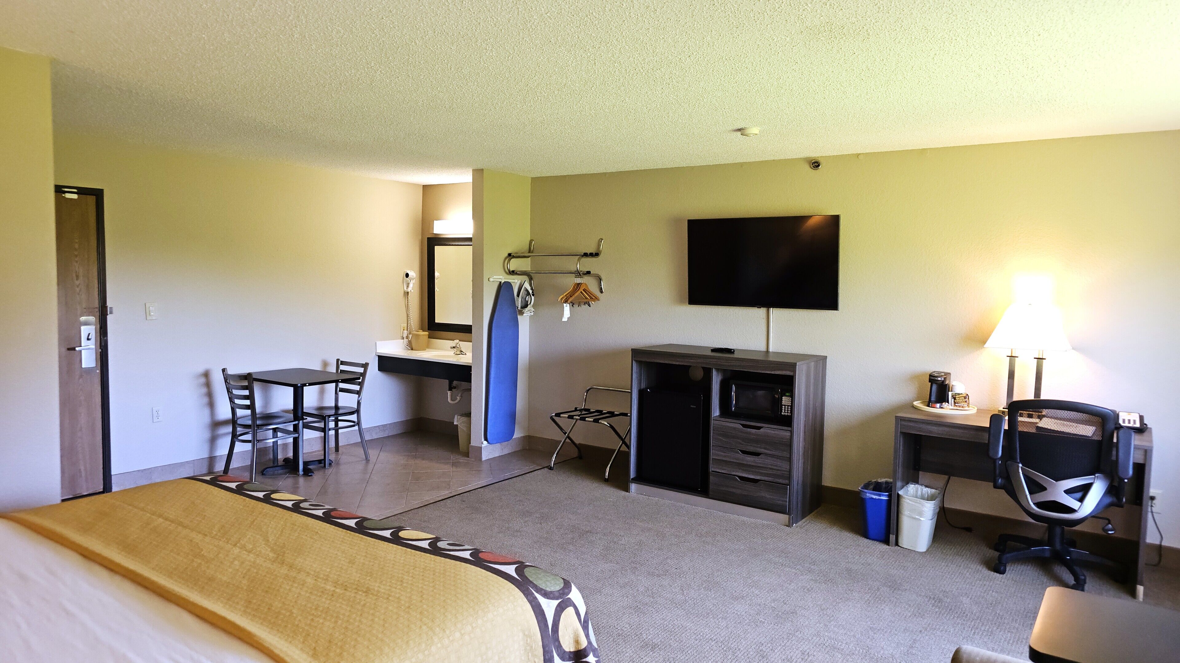 A hotel room with a bed, a desk, a chair, a TV, a lamp, and a microwave.