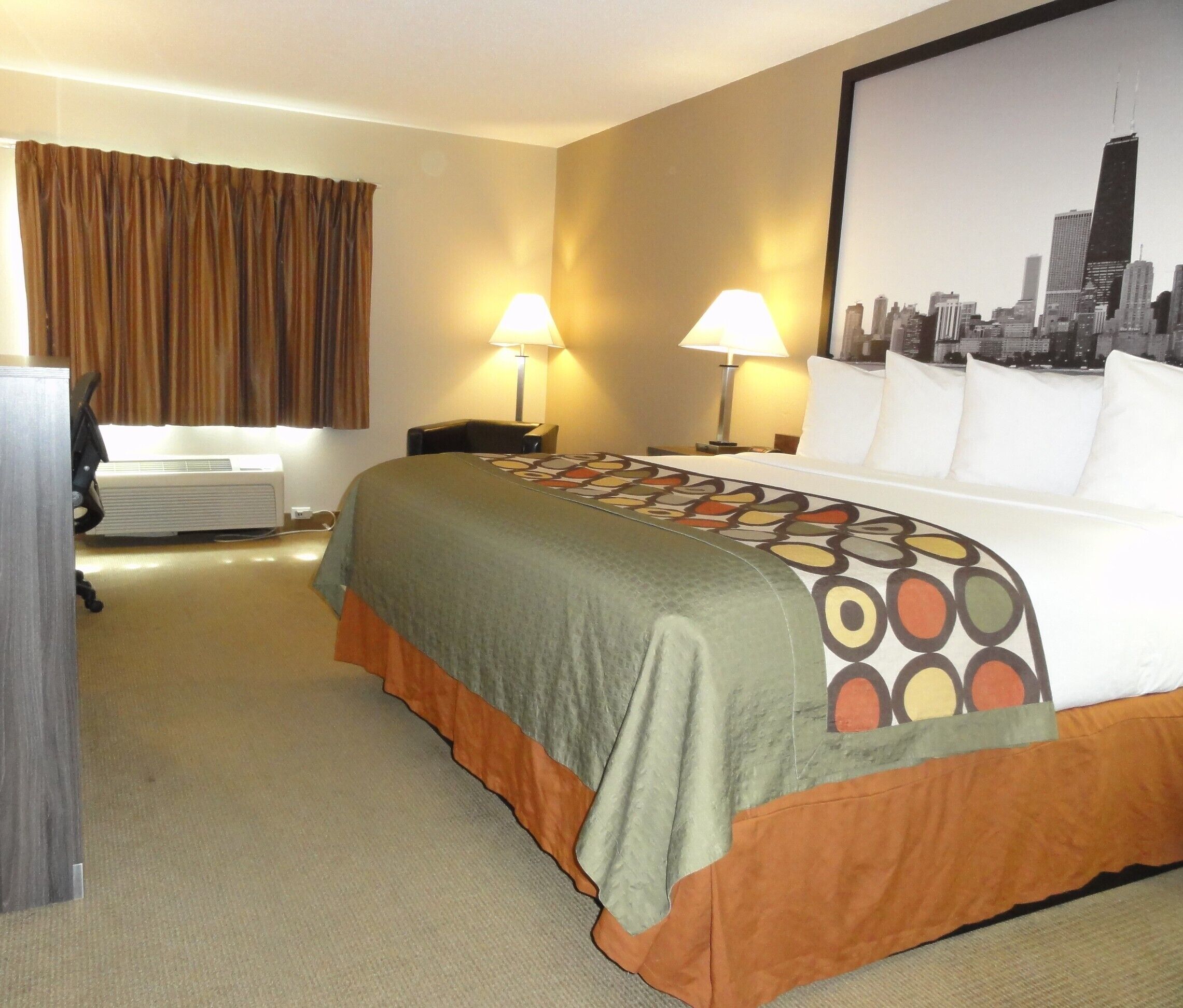 A hotel room with a large bed, two bedside lamps, a desk with a chair, an air conditioning unit, and a cityscape poster on the wall.