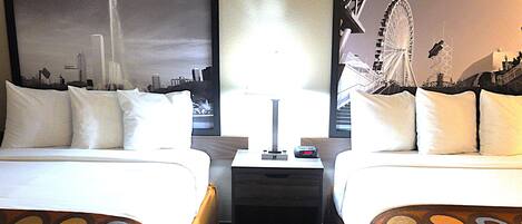 A hotel room with two beds, a nightstand, a lamp, and two framed pictures of cityscapes.