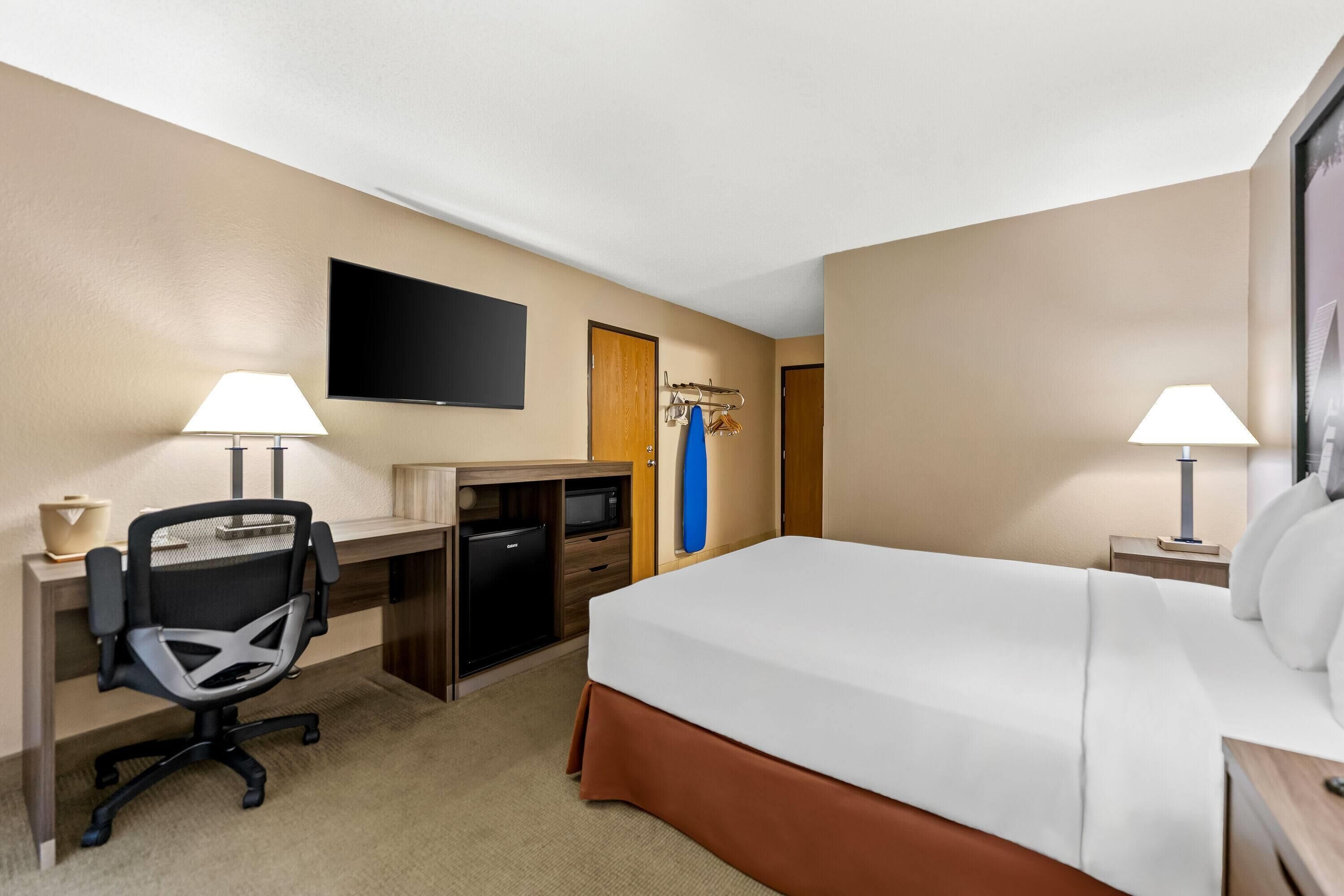 A hotel room with a bed, desk, chair, TV, and a small kitchenette.
