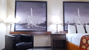 Habitación de hotel con una cama, un sillón de cuero negro, una mesita de noche con una lámpara y dos cuadros grandes con vistas a un paisaje urbano que incluye una fuente.