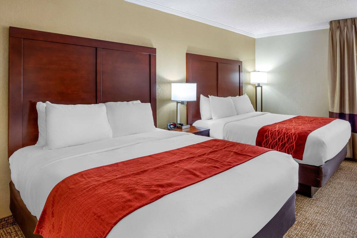 Habitación de hotel con dos camas, cada una con ropa de cama blanca y mantas rojas. Hay mesitas de noche con lámparas y un reloj. La habitación tiene un cabecero de madera y suelo alfombrado.